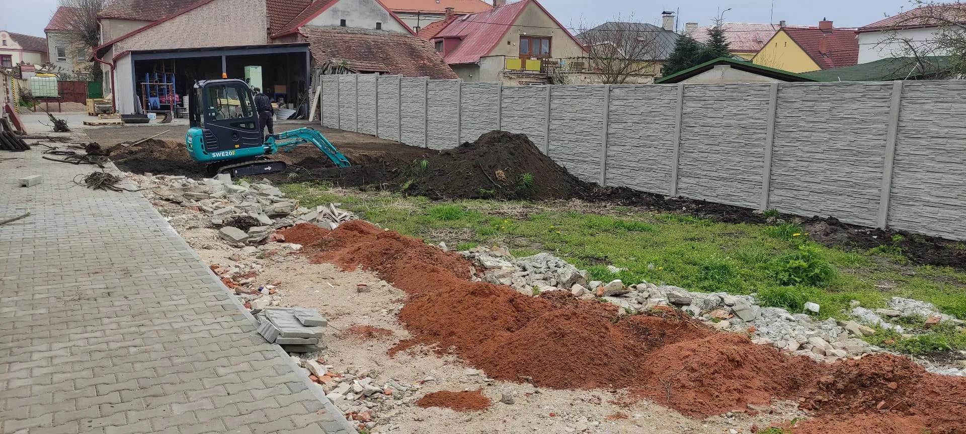 Minibagr při úpravě terénu zahrady podél nového betonového plotu
